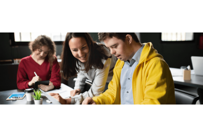 a female educator and male student, looking down at what appears to be a paper or project on a school desk. The educator is smiling, and the student is focused on the paper. 