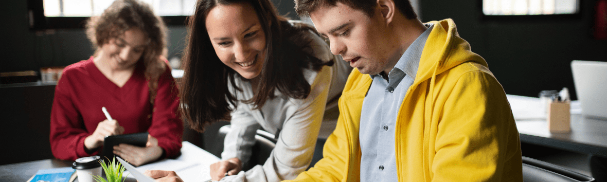 a female educator and male student, looking down at what appears to be a paper or project on a school desk. The educator is smiling, and the student is focused on the paper. 