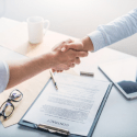 Two people shaking hands over a desk with a document, glasses, a pen, and a coffee cup present