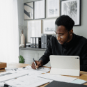 A man with glasses in an office sits in front of a desk writing holding a pen on top of a paper while keeping other documents and a table on top of the desk